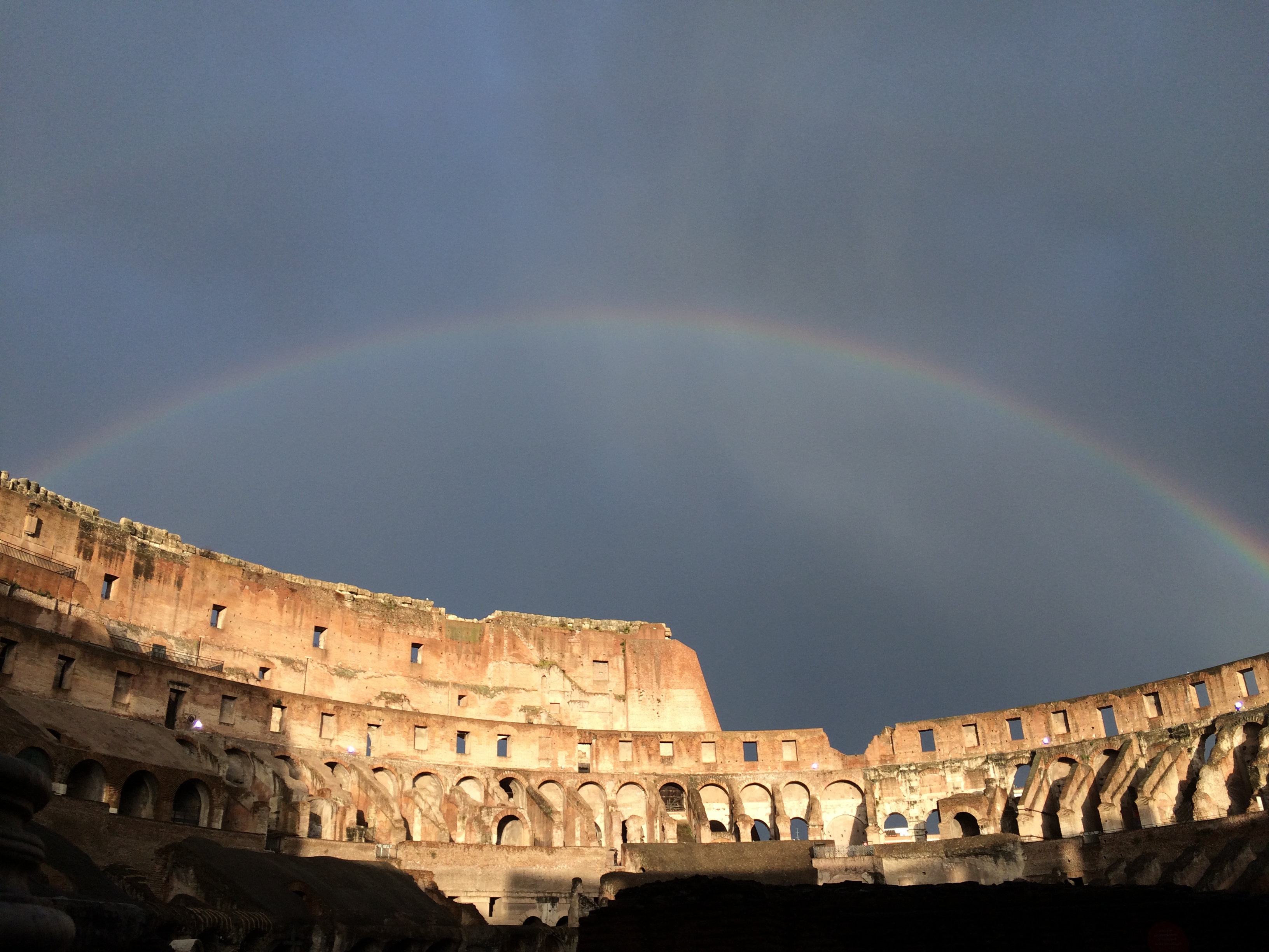Rainbow above the Colosseum in Rome.  Sunshine and shadow in the foreground.  Cloud vapour streaking the sky.